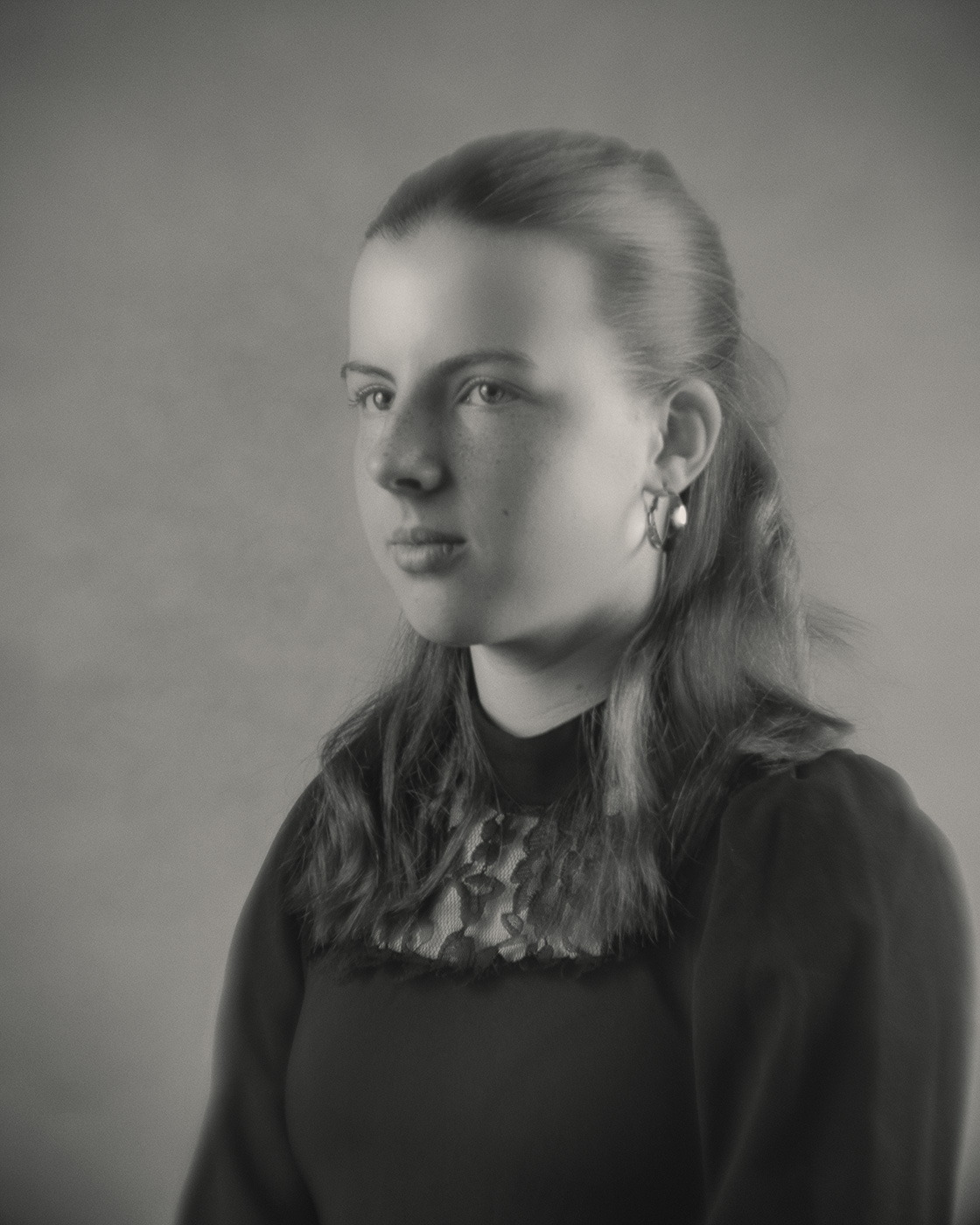 Ein zwölfjähriges Mädchen mit mittellangem, nach hinten gekämmtem Haar blickt leicht zur Seite. Sie trägt ein dunkles Oberteil mit einem auffälligen, gemusterten Kragen und Ohrringe. Das Bild wirkt durch das Weichzeichner-Objektiv sanft und hat einen nostalgischen Charakter.