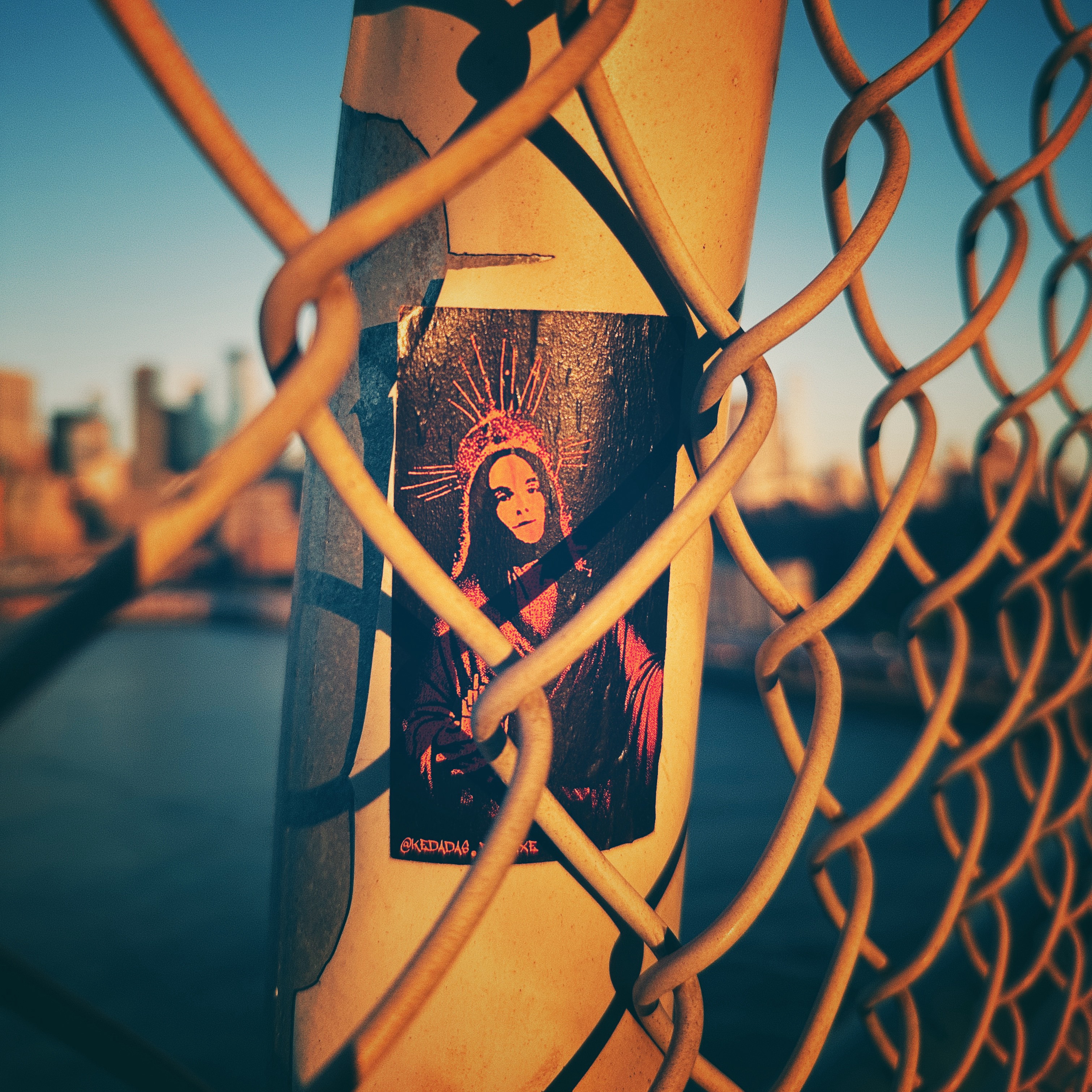 Sunrise light hitting a sticker of a saint on the Manhattan Bridge fence