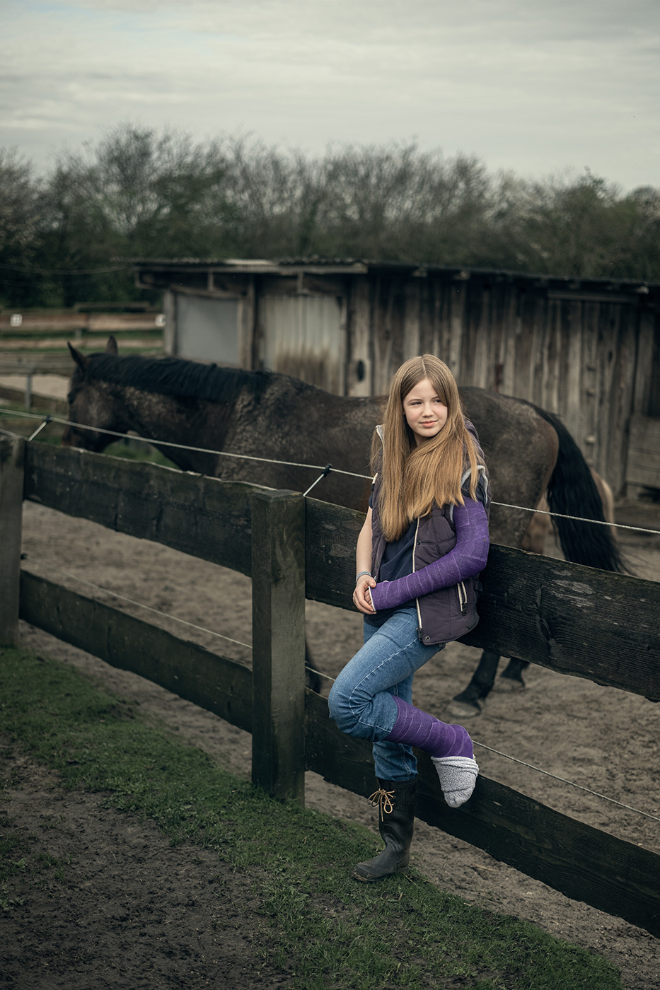 Ein 12-jähriges Mädchen mit langem, offenem Haar lehnt an einem Holzzaun. Ihr linkes Bein und ihr linker Arm sind in Gips, beide Gipse sind violett. Sie trägt eine blaue Jeans, eine dunkle Jacke mit lilafarbenen Ärmeln und einen schwarzen Gummistiefel. Die Zehen, die aus dem Gipsbein rausschauen würden, sind von einer grauen Socke verdeckt. Im Hintergrund steht ein dunkles Pferd vor einem hölzernen Stallgebäude. Die Szene spielt auf einem matschigen, ländlichen Gelände unter bewölktem Himmel.